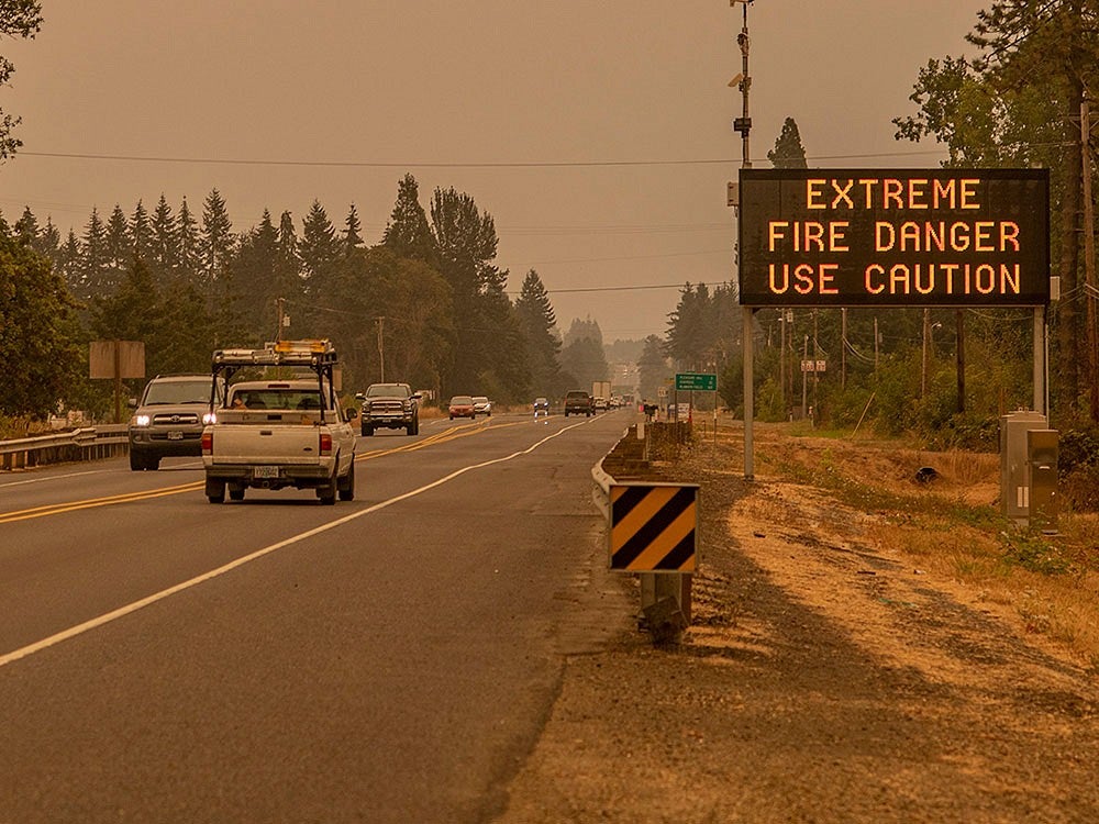 Tan-colored sky with heavy smoke obscures visibility on a busy highway lined with trees. Cars drive by a sign that warns ”Extreme Fire Danger Use Caution”.