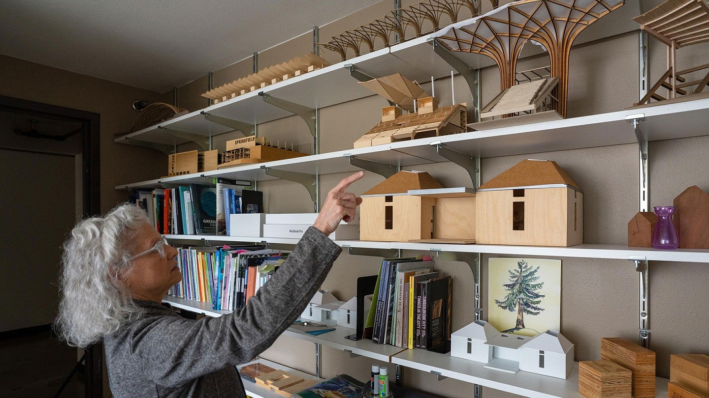 Professor Judith Sheine points at wooden building models displayed on a shelf