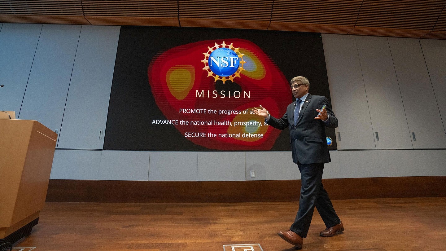 During his visit to the UO Eugene campus, NSF director Sethuraman Panchanathan strides to the podium in front of a screen that reads "NSF mission: promote the progress of science, advance the national health, prosperity and welfare. Secure the national defense."