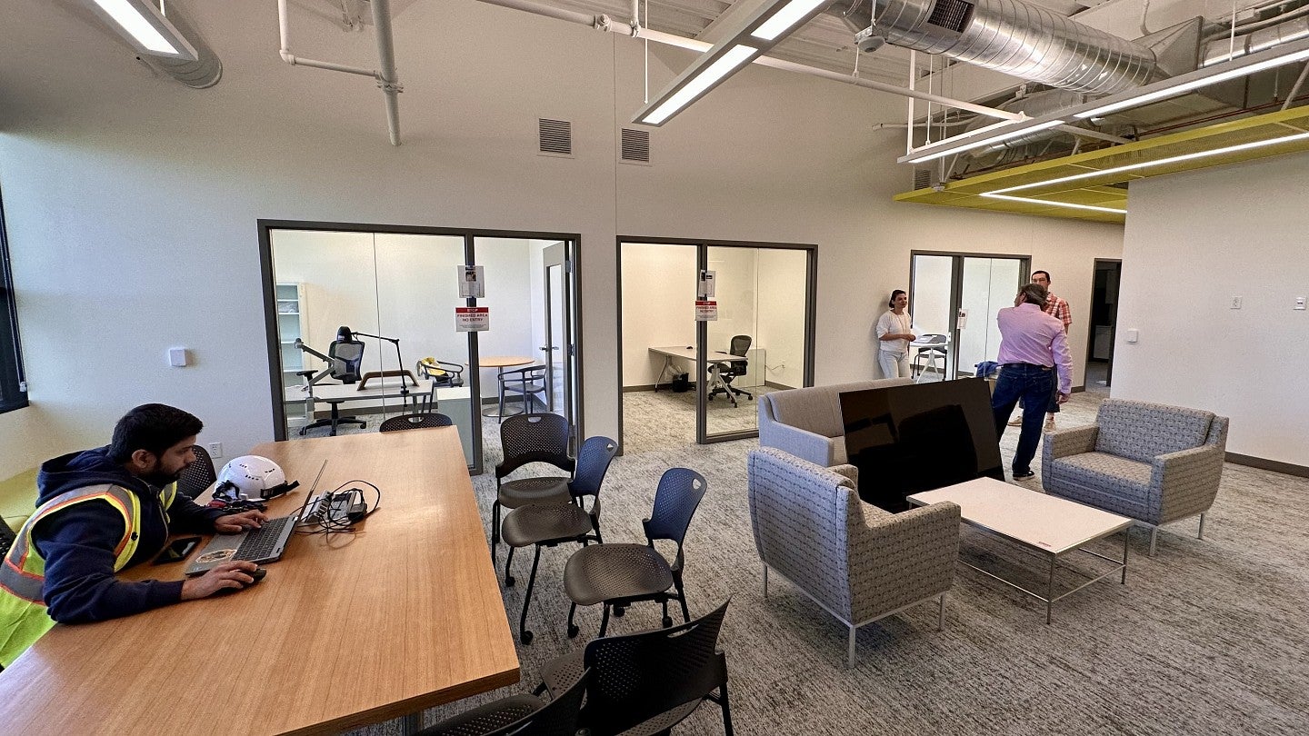 UO Portland Innovation Building open office space with an employee sitting at a table working on a computer and other employees across the room standing and talking behind a loveseat and chairs