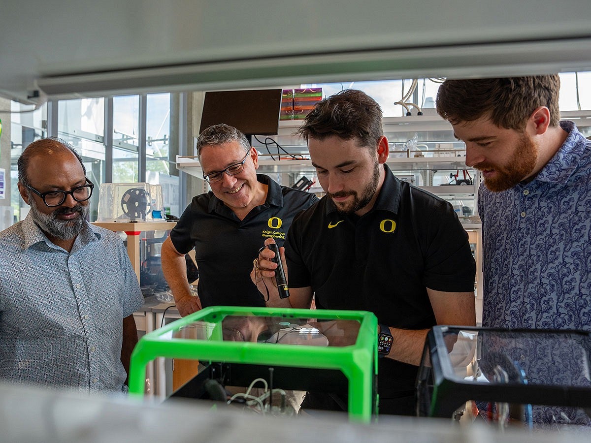 Four scientists in a lab shine a UV flashlights onto a glowing material near a 3D printer