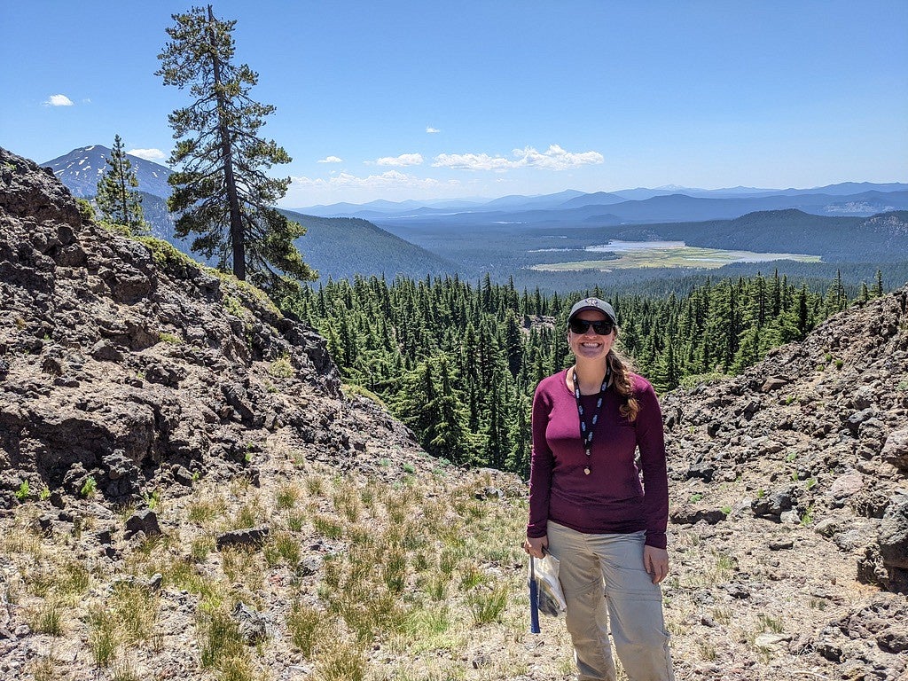 Annika Dechert, in a magenta jacket, stands on a rocky slope with mountains and lakes in the background.