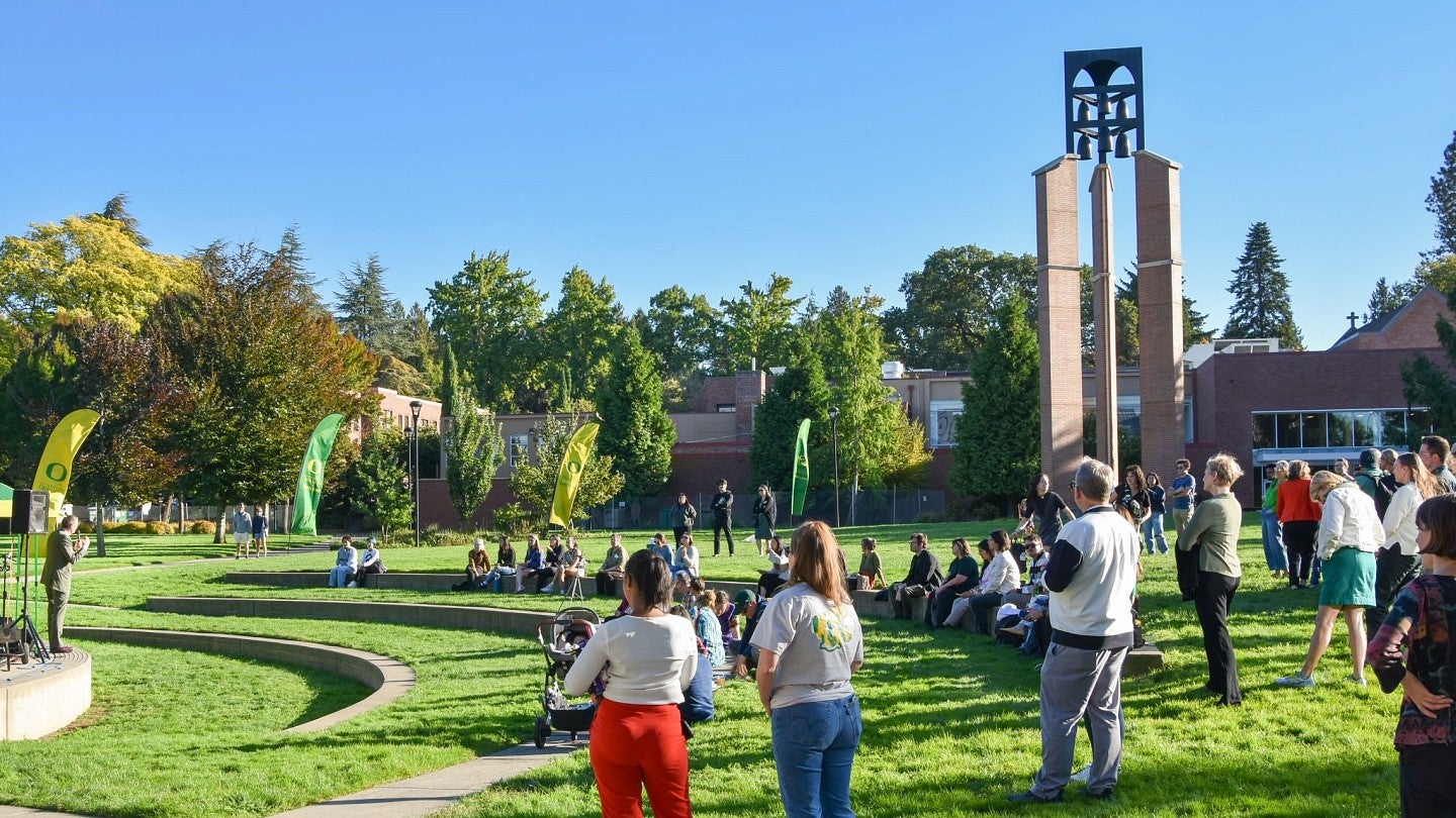 Uo community members gathered in open grass space listening to President Scholz speak from a stage with campus buildings in the background