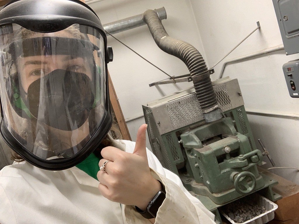 Annika Dechert gives a thumbs up while wearing a face shield and standing next to a metal machine that is crushing rocks.