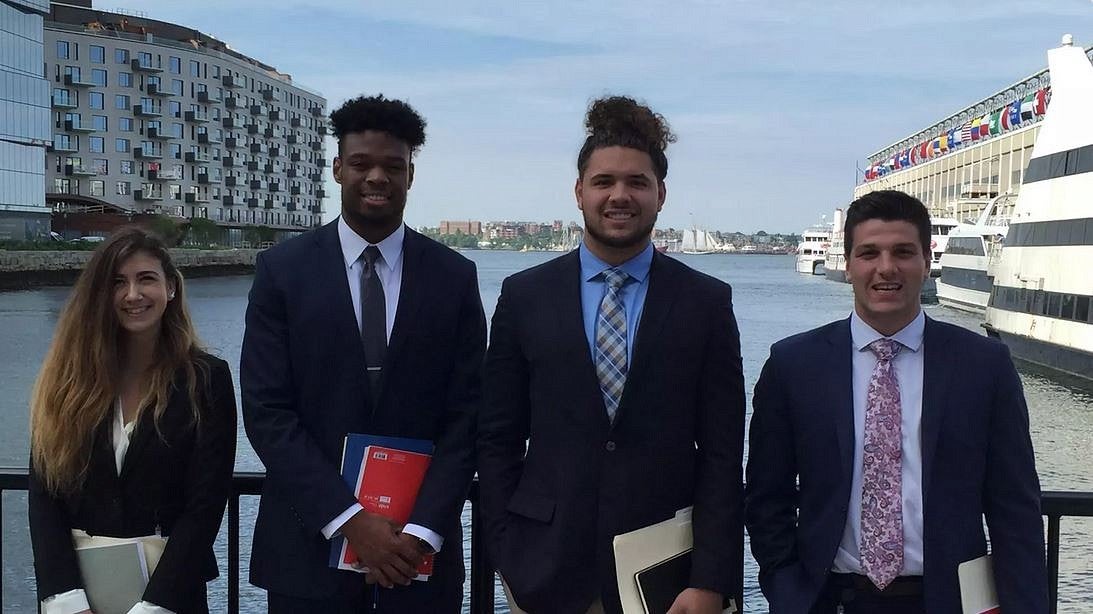 Distance runner Melissa Berry and three football players — Gus Cumberlander, Cam McCormick and Ben Gomes wearing business suits, standing at a waterfront with urban landscape in background.