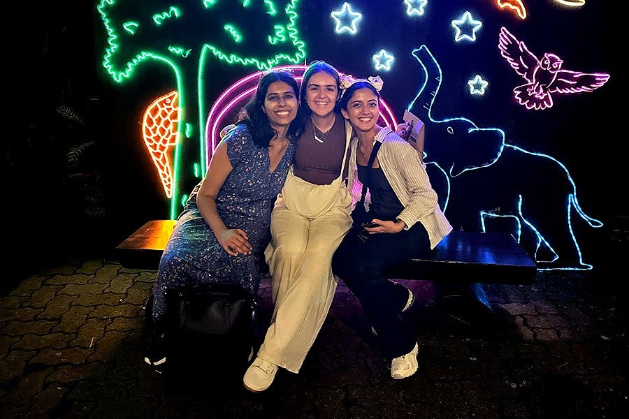 Three smiling young women sitting on a bench, colorful neon signage in the background depicts a tree, elephant, owl, pangolin. and stars