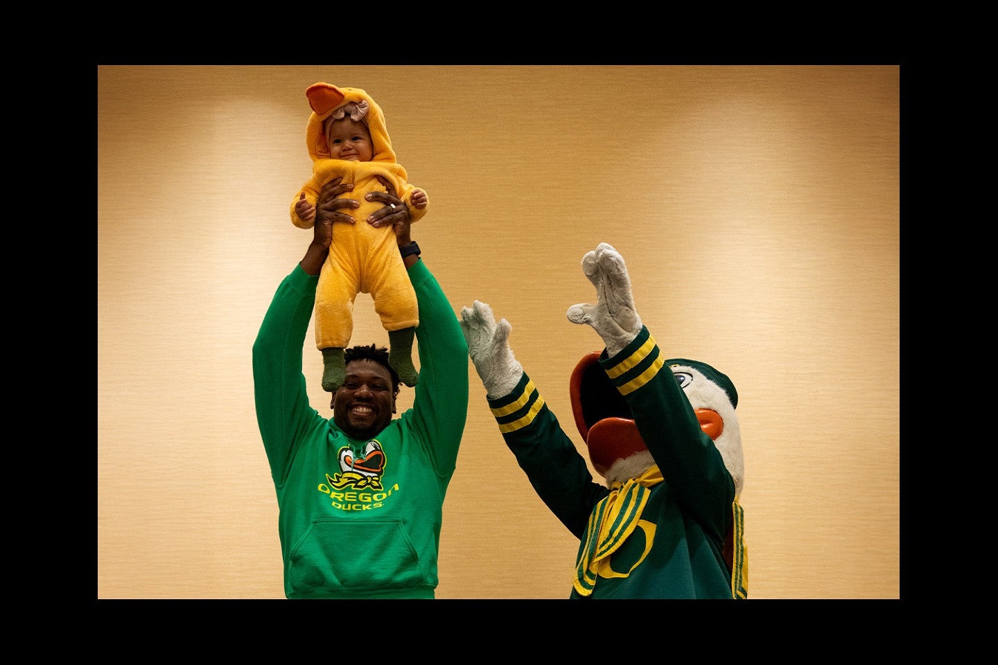 a small child in a duck costume is held aloft by her father while the Duck also reaches up