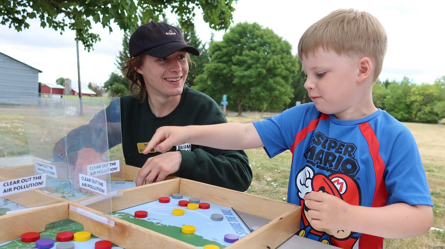 A museum outreach educator smiles as a boy points to a spot on a board game about protecting the environment