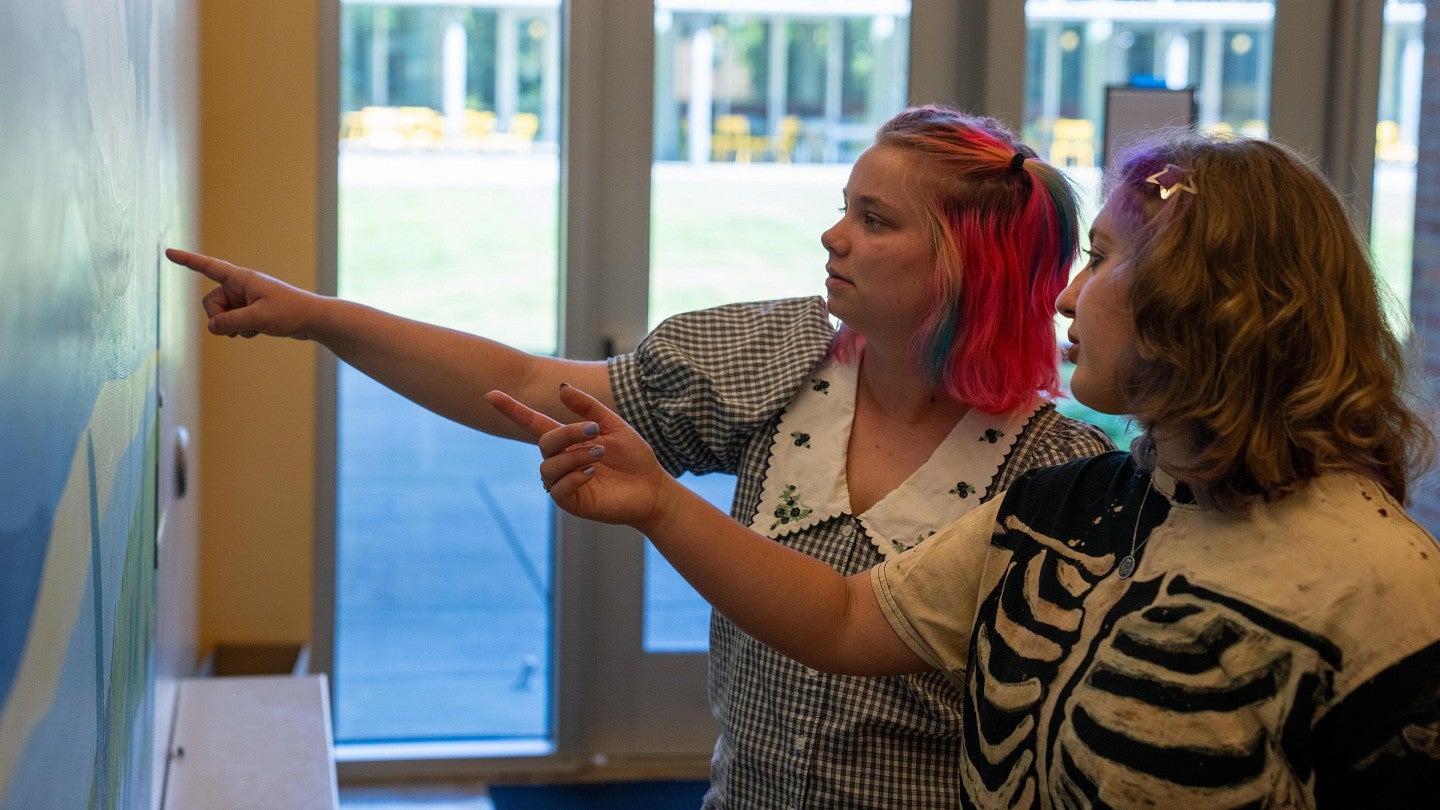 Two student artists point at a detail on their mural