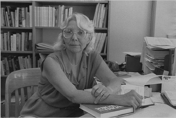 Joan Acker sitting at her office desk, hand resting on a book