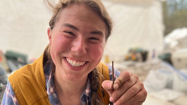 Kelby Beyer, a young woman wearing a yellow work vest and checkered shirt, holds up a bone needle.