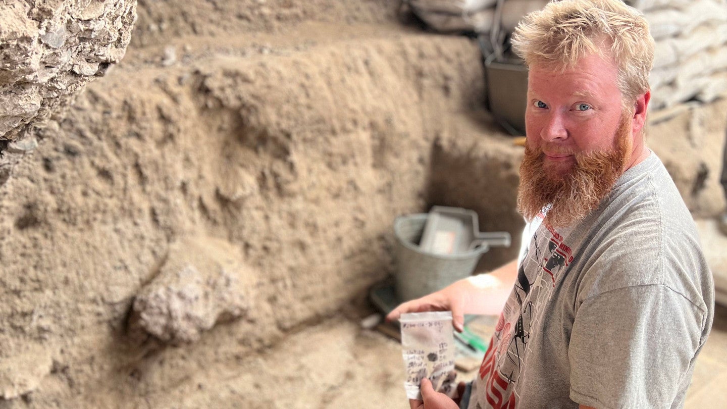 Matt Skeels, a young man with a beard and gray shirt, is holding a bag with an obsidian tool inside.