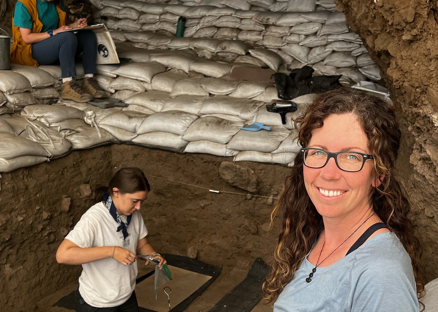 Katelyn McDonough, anthropologist, stands near an excavation site with students digging in background.