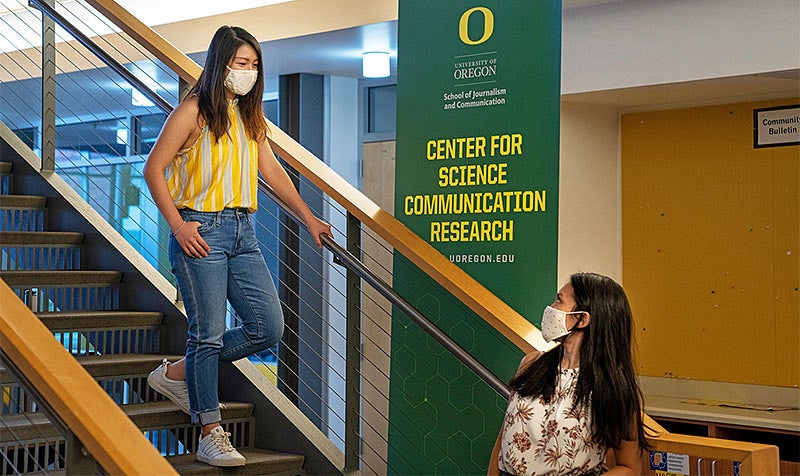 A student wearing a mask walking down the stairs toward another student wearing a mask in Allen Hall with the SOJC banner behind them