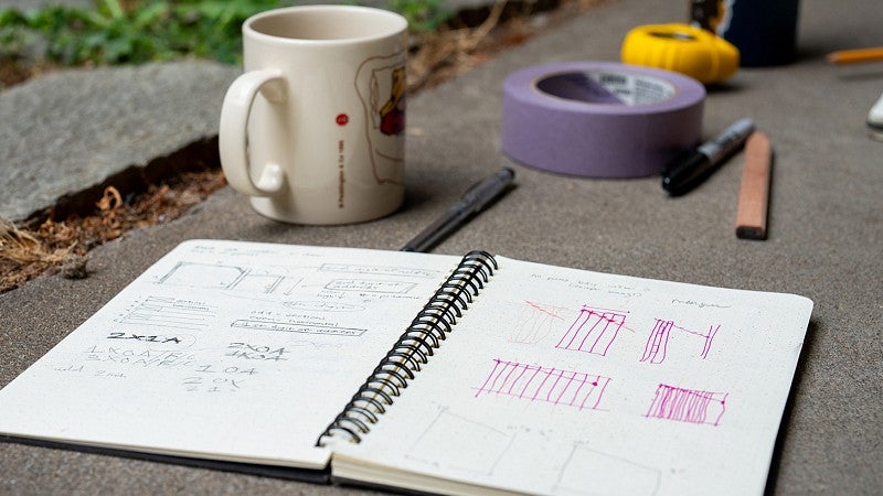 A coffee cup, tape measure, tape, pencil, water bottle, and open note pads with design sketches during the Bruton Design Intensive