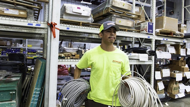 A young man in a yellow T-shirt and cap with a company logo carries electrical cables.