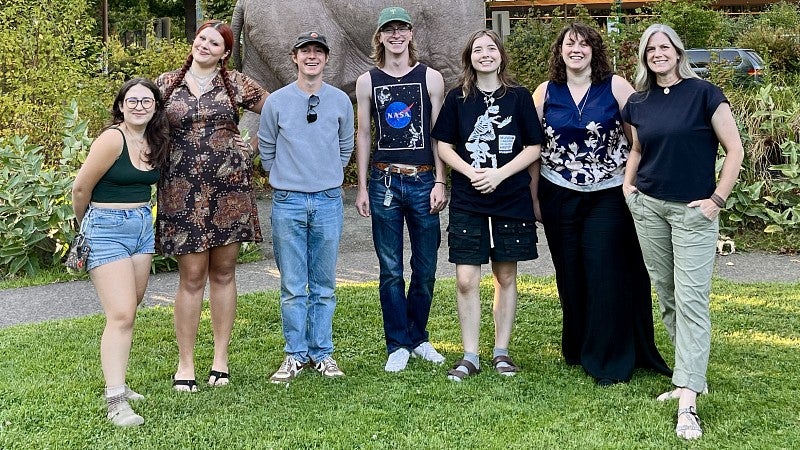 Student outreach educators and staff of the Museum of Natural and Cultural History pose in front of a statue of an elephant at the museum