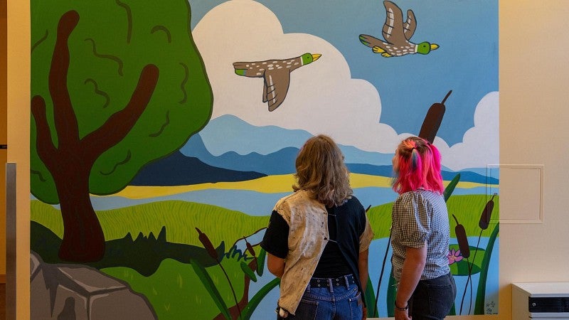 Two student artists stand in front of their mural, which depicts trees, ducks and a mountainous horizon