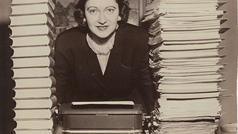 Peg Lynch with typewriter and tall stacks of scripts.