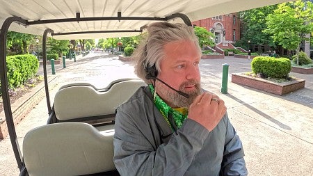 A man drives a golf cart on the UO campus while speaking into a headset