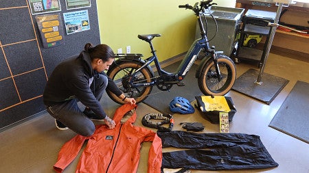 A bike shop manager arranges bike gear including rain gear, charger and pannier, next to an electric bike