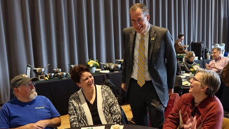 A man stands before a table with people sitting, all chatting and smiling