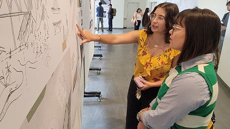 A student gestures to a wall-sized design of an airport while an observer listens