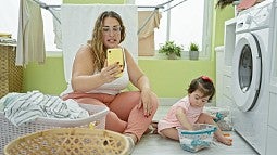 A parent doing laundry with a child whild looking at cellphone