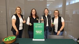 Four women from the UO Benefits Office each wearing a black vest and holding a rubber duck in their right hand standing behind a table at a benefits fair