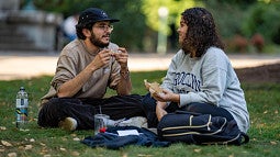 two students chat while sitting cross-legged on a lawn and eating