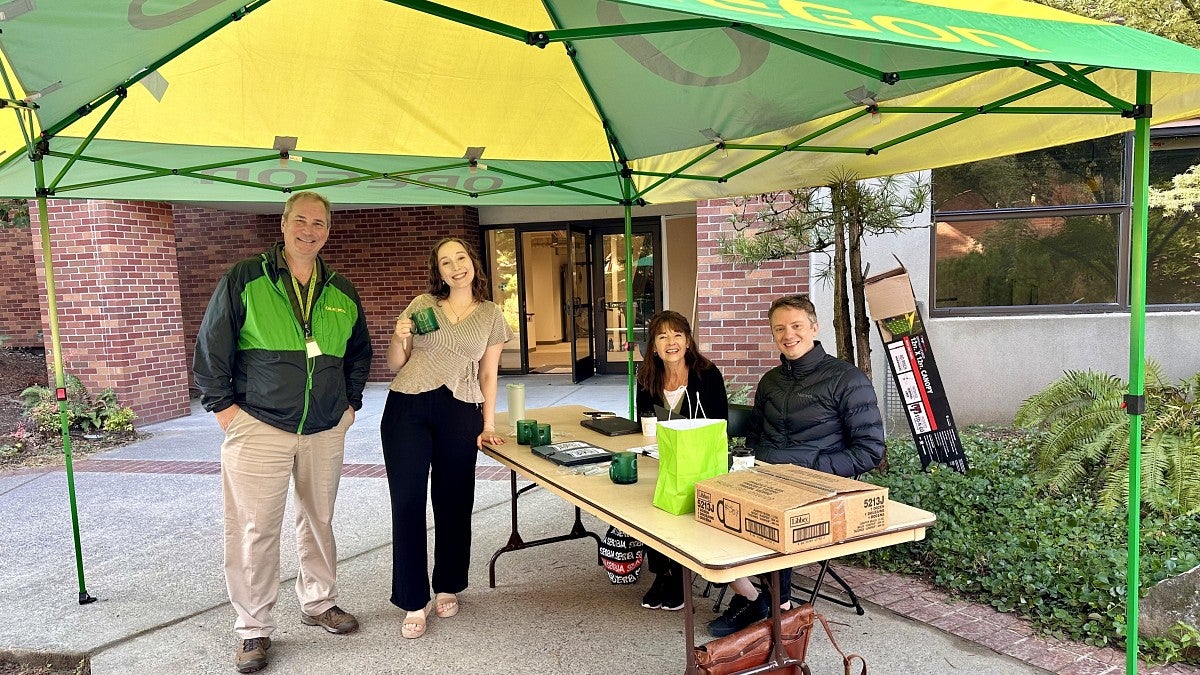 UO employees standing and sitting at a table under a green and yellow Oregon canopy welcoming others to the Northeast Portland campus 