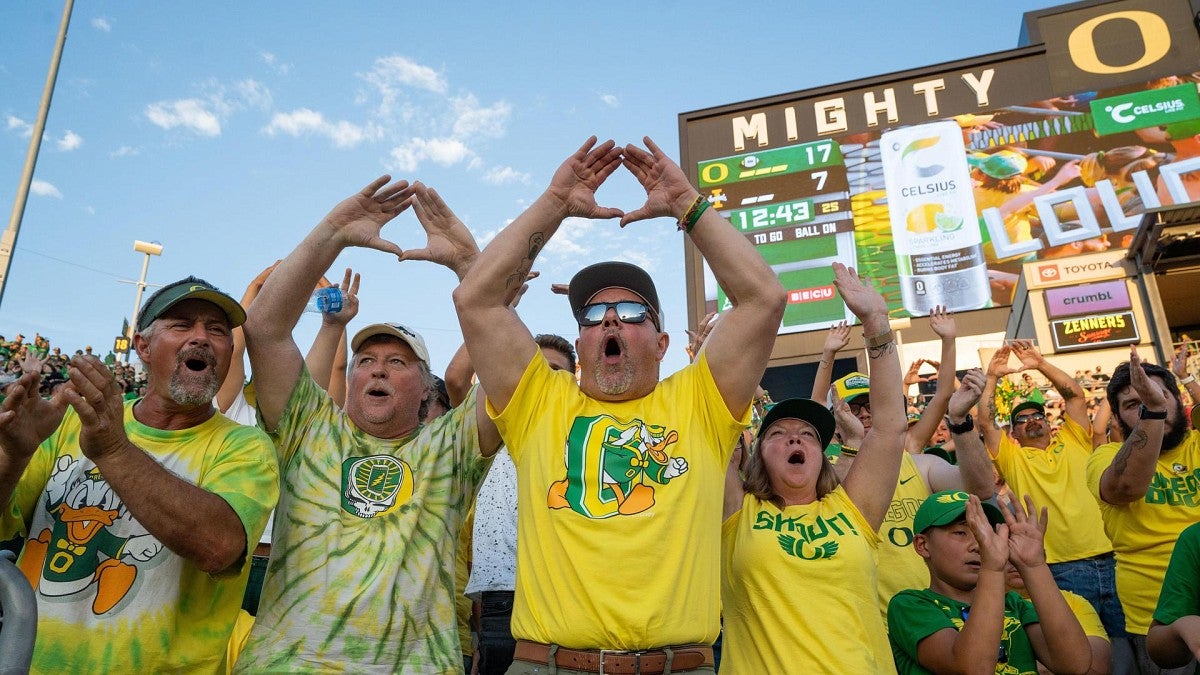 Oregon Duck fans dressed in green and yellow raising their hands overhead to make an O as they cheer at a football game. 