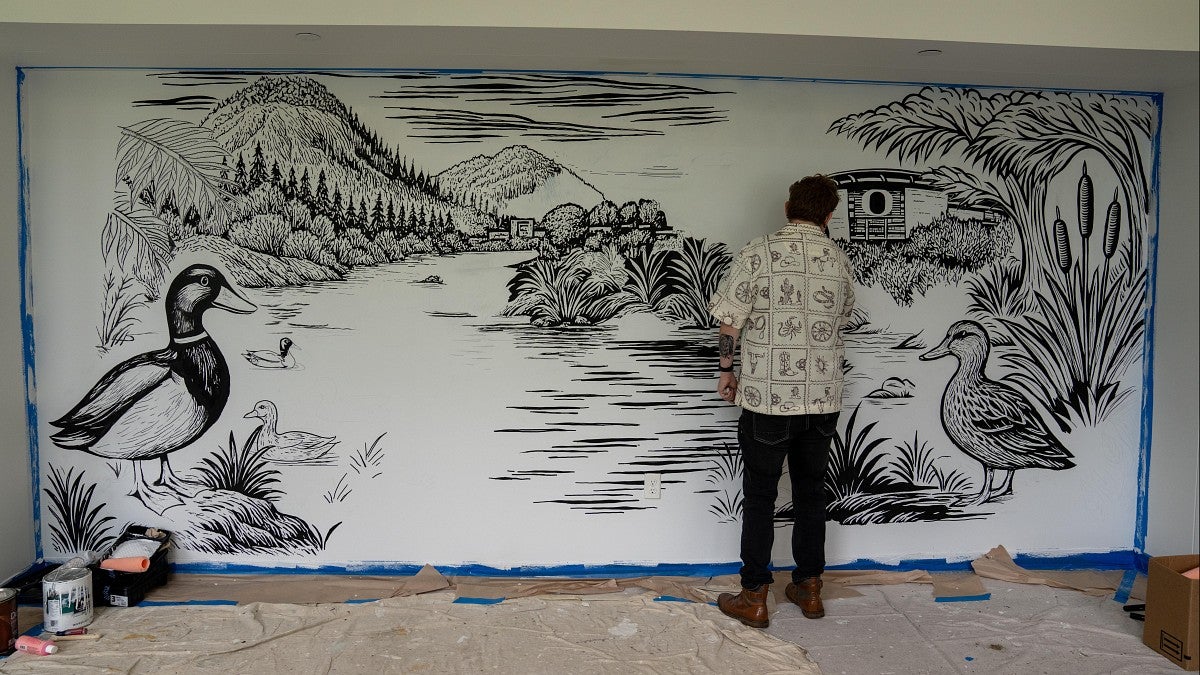 A man uses a marker to draw a black-and-white mural of birds, mountains and a river