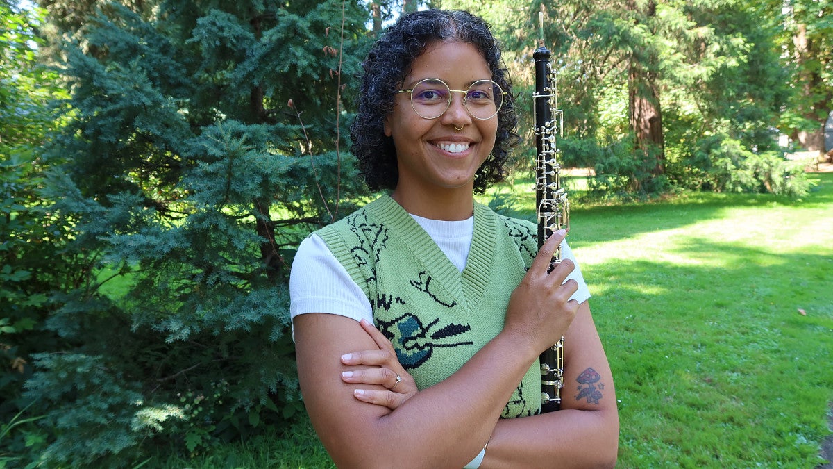 A woman smiles while holding an oboe in a tree-filled outdoor setting