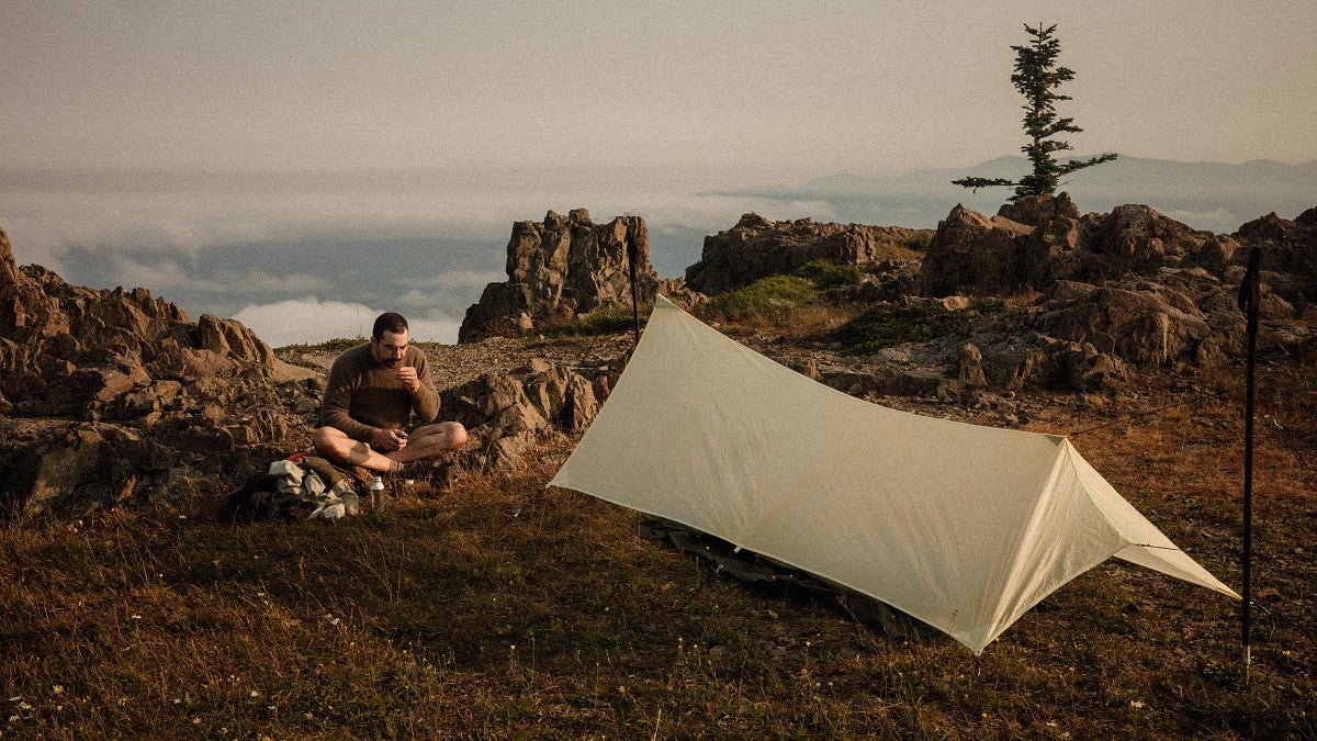 A person drinks next to a tent in a picturesque coastal setting