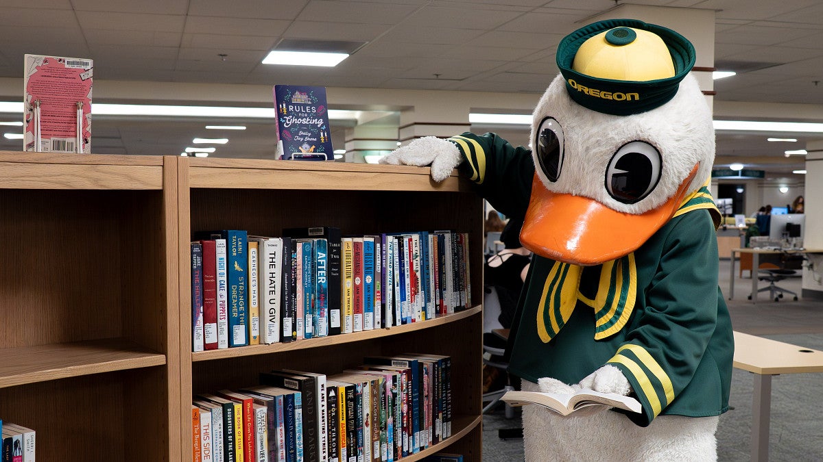The Duck reads next to a bookshelf in the Knight Library