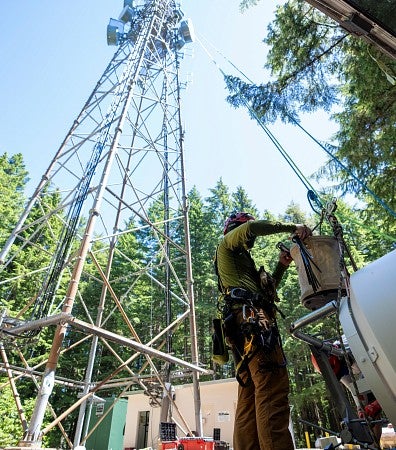 A man in climbing gear prepares a microwave antenna to be hoisted up a tower