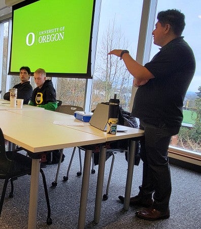 A man speaks to students in a classroom setting with a large screen in the background