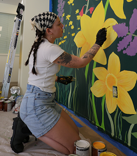 An artist kneels while painting a flower on a mural
