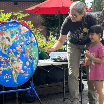 A woman standing with a child points to a board game that features a depiction of the Earth