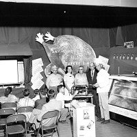 Exhibitors and state fair attendees in front of turkey-shaped sign.